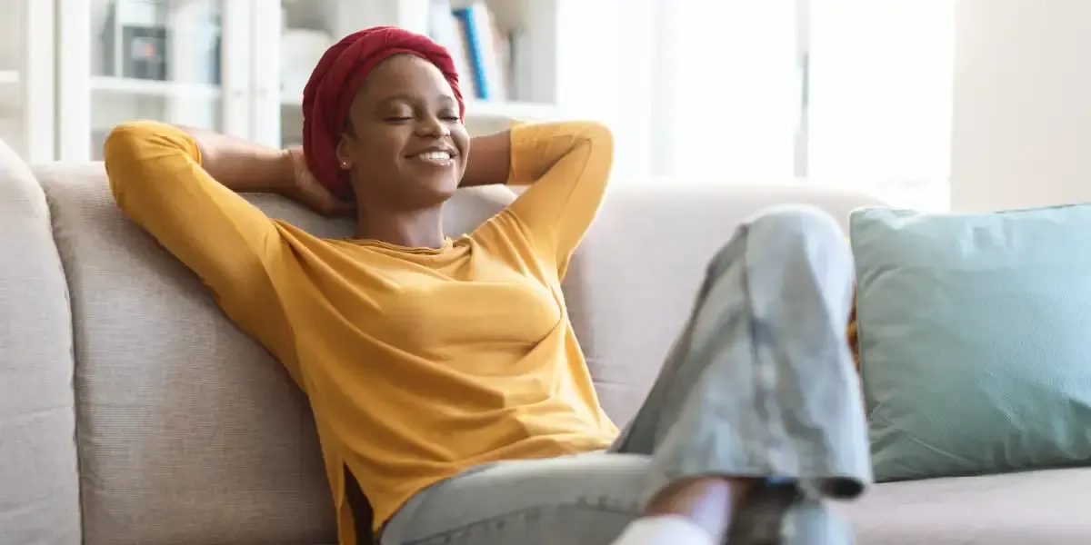 A woman sitting on a couch with her hands behind her head, her eyes closed, smiling