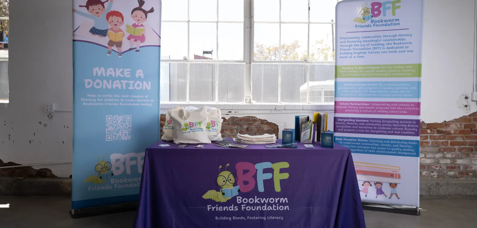 Two vinyl banners and a table with a tablecloth contains tote bags and printed materials, all branded with Bookworm Friends Foundation (BFF).