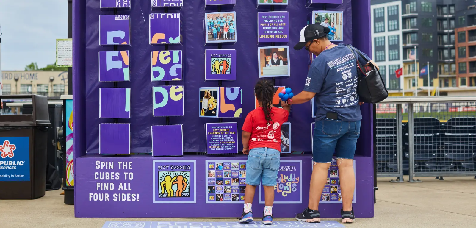 Small girl and adult stand side-by-side in front of a large display of rotating blocks, spinning the blocks to reveal facts about Best Buddies.