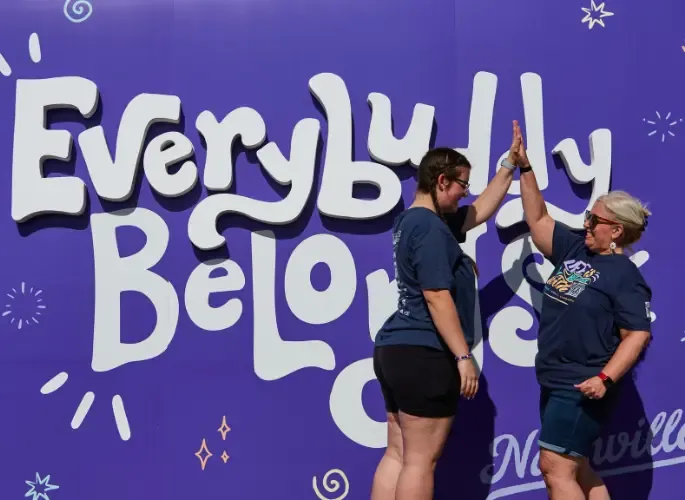 Two women high-five in front of a large purple sign with a standout portion that says 