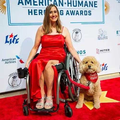 A woman in a wheelchair next to a small dog on a red carpet with gold stars and in front of a backdrop with sponsor logos.