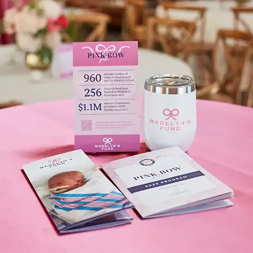 A Madelyn's Fund brochure, program, table tent, and tumbler on top of a table with a pink tablecloth