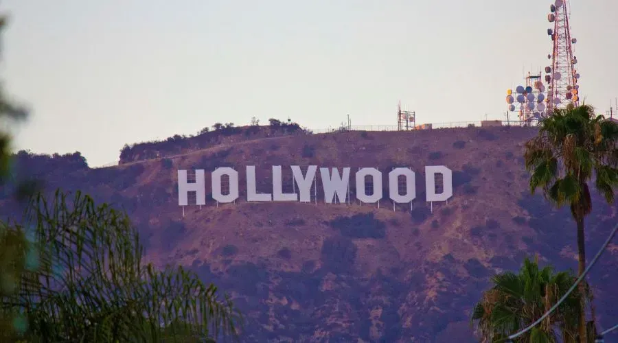 A close-up view of the iconic Hollywood sign situated on the hills of Los Angeles, California. The large white letters spelling “HOLLYWOOD” are set against a backdrop of dry brown hills with some greenery in the foreground. Communication towers can be seen on the hilltop next to the sign, highlighting the famous landmark that represents the entertainment industry.